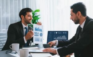 two men suits sit desk one them is holding graph other is showing graph